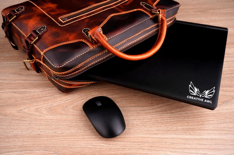 May include: A brown leather briefcase with a laptop computer inside. The laptop has a white owl logo with the text "CREATIVE AWl" on the back. A black wireless mouse is on a wooden surface.