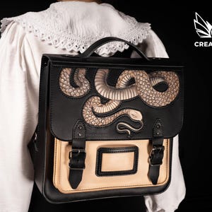 Könnte beinhalten: Schwarzer und hellbrauner Lederrucksack mit detailliertem Schlangenmuster auf der Klappe. Die Tasche hat einen oberen Griff, verstellbare Träger und eine rechteckige Vordertasche mit Schnallenverschlüssen. Das Creative Awl-Logo ist sichtbar.
