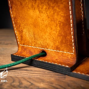 May include: Close-up of a handcrafted leather phone stand with a rich, warm brown finish. The stand features white stitching and a green cord passing through a hole. The Creative Awl logo is visible at the bottom.