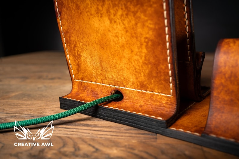 May include: Close-up of a handcrafted leather phone stand with a rich, warm brown finish. The stand features white stitching and a green cord passing through a hole. The Creative Awl logo is visible at the bottom.