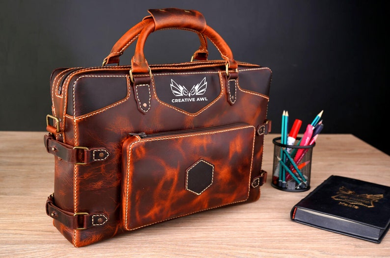 May include: A brown leather briefcase with a front pocket and a zippered compartment. The briefcase has a top handle and a buckle closure. The briefcase has the text "CREATIVE AWL" and a logo of a bird with wings spread.