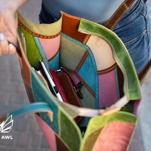 May include: A multi-coloured leather tote bag featuring patchwork panels in shades of green, pink, blue, and brown. The interior is visible, containing a small red pouch and a black notebook. The bag has stitched seams and thin leather straps.