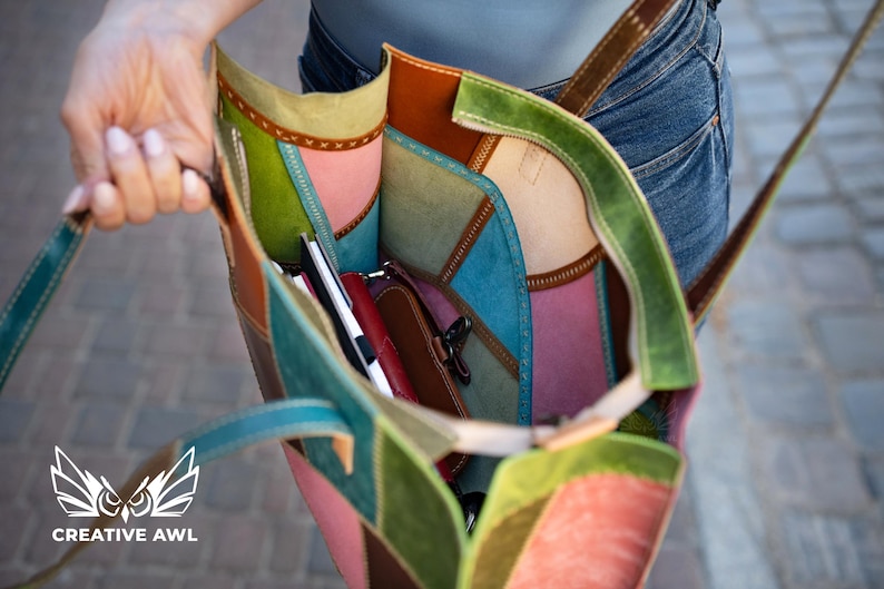 May include: A multi-coloured leather tote bag featuring patchwork panels in shades of green, pink, blue, and brown. The interior is visible, containing a small red pouch and a black notebook. The bag has stitched seams and thin leather straps.