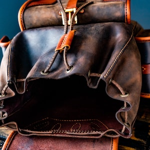 May include: A brown leather backpack with a drawstring closure and a flap with a buckle. The backpack is open and the inside is visible.
