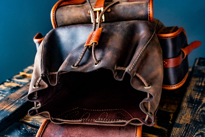 May include: A brown leather backpack with a drawstring closure and a flap with a buckle. The backpack is open and the inside is visible.