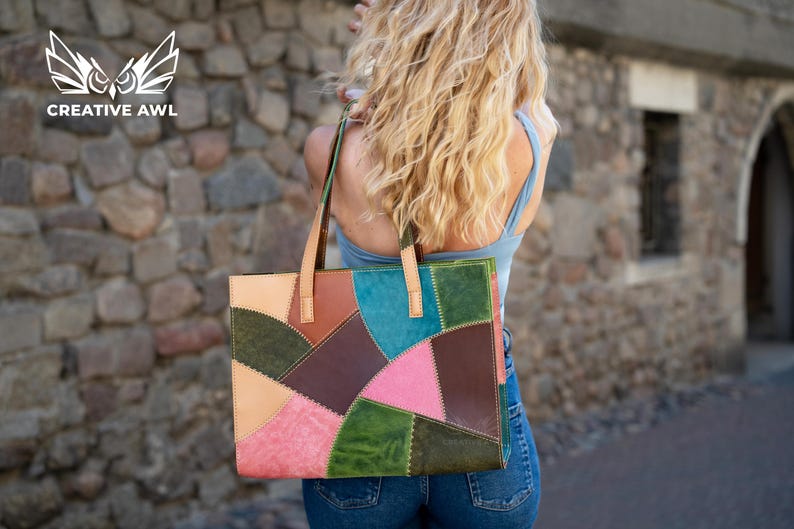 May include: A rectangular tote bag constructed from multi-coloured leather patches in shades of pink, green, brown, and blue. The bag features two light tan leather shoulder straps and visible stitching along the edges of each leather panel.