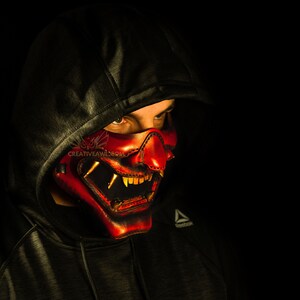 K&ouml;nnte beinhalten: Eine Person tr&auml;gt einen schwarzen Kapuzenpullover mit einer roten und goldenen Oni-Maske, die ihr Gesicht bedeckt. Die Maske hat scharfe Z&auml;hne und H&ouml;rner.