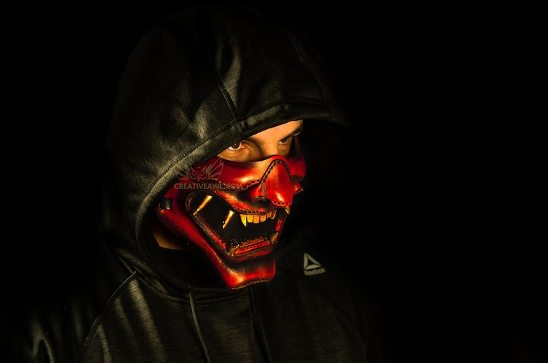 K&ouml;nnte beinhalten: Eine Person tr&auml;gt einen schwarzen Kapuzenpullover mit einer roten und goldenen Oni-Maske, die ihr Gesicht bedeckt. Die Maske hat scharfe Z&auml;hne und H&ouml;rner.