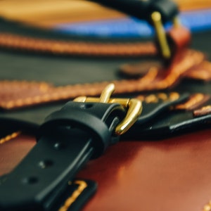May include: Close-up of a leather bag with a black strap and a gold-colored buckle. The bag has a brown and black color scheme with decorative stitching. The image highlights the craftsmanship and detail of the bag's design.