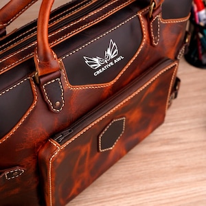 May include: A brown leather briefcase with a zippered pocket and a front pocket with the text "CREATIVE AWL" in silver lettering. The briefcase has a top handle and a shoulder strap.