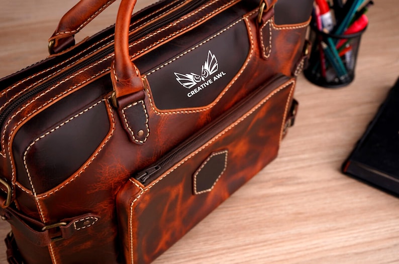 May include: A brown leather briefcase with a zippered pocket and a front pocket with the text "CREATIVE AWL" in silver lettering. The briefcase has a top handle and a shoulder strap.