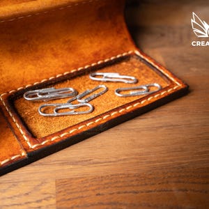 May include: Close-up of a handcrafted leather wallet with a compartment holding several silver paper clips. The wallet is a warm brown color with visible stitching and a dark border. The Creative Awl logo is in the upper right corner.