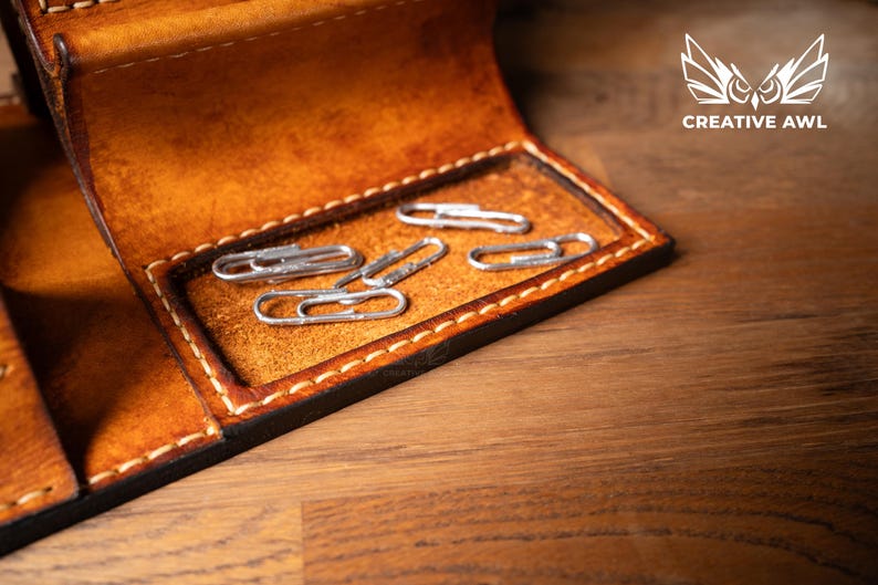 May include: Close-up of a handcrafted leather wallet with a compartment holding several silver paper clips. The wallet is a warm brown color with visible stitching and a dark border. The Creative Awl logo is in the upper right corner.