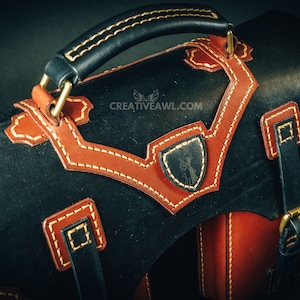 May include: Black and brown leather satchel with tan stitching and accents. Features a top handle, shoulder strap, and decorative shield emblem. The bag has a classic design with brass hardware.