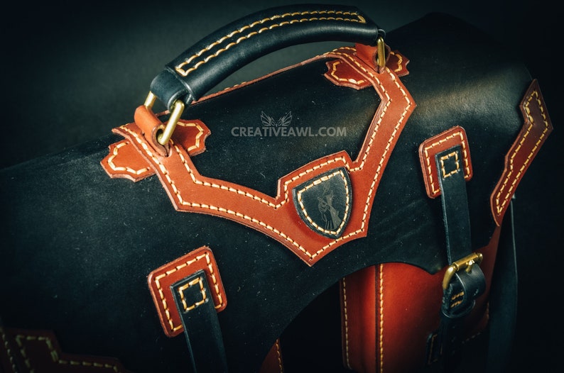 May include: Black and brown leather satchel with tan stitching and accents. Features a top handle, shoulder strap, and decorative shield emblem. The bag has a classic design with brass hardware.