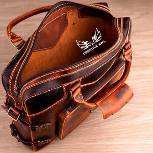 May include: A brown leather briefcase with a zippered closure and multiple pockets. The interior is lined with a tan fabric and features the logo "CREATIVE AWL" in white.