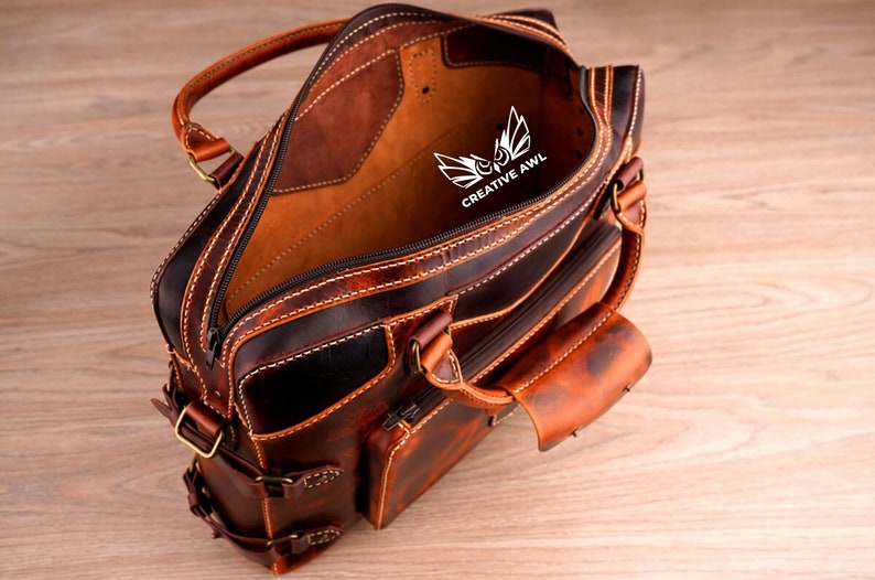 May include: A brown leather briefcase with a zippered closure and multiple pockets. The interior is lined with a tan fabric and features the logo "CREATIVE AWL" in white.