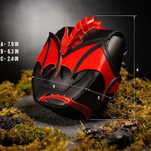 Op de afbeelding: Een zwarte en rode tas met draakthema, voorzien van een gebeeldhouwde drakenkop en vleugels op de voorflap. De afmetingen zijn aangegeven als A: 20 cm, B: 16 cm en C: 6 cm. De tas heeft een textuurafwerking en een puntig staartdetail onderaan.