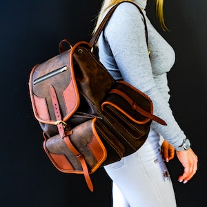 May include: A brown leather backpack with a large front pocket and a smaller pocket on the side. The backpack has a buckle closure and a zipped pocket on the top.