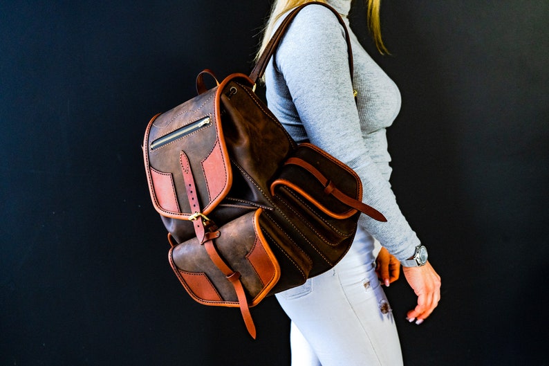 May include: A brown leather backpack with a large front pocket and a smaller pocket on the side. The backpack has a buckle closure and a zipped pocket on the top.