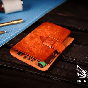 May include: A brown leather tool roll with a flap closure and a decorative stitching detail. The roll is open and contains various tools, including awls, punches, and a green marker.