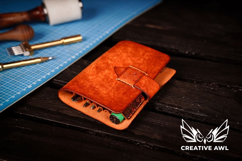 May include: A brown leather tool roll with a flap closure and a decorative stitching detail. The roll is open and contains various tools, including awls, punches, and a green marker.