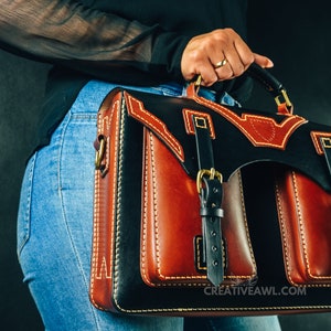 May include: A handcrafted leather briefcase with a striking design. The bag features a combination of black and brown leather, accented with cream stitching and gold-colored hardware. The bag has two front pockets and a top handle.