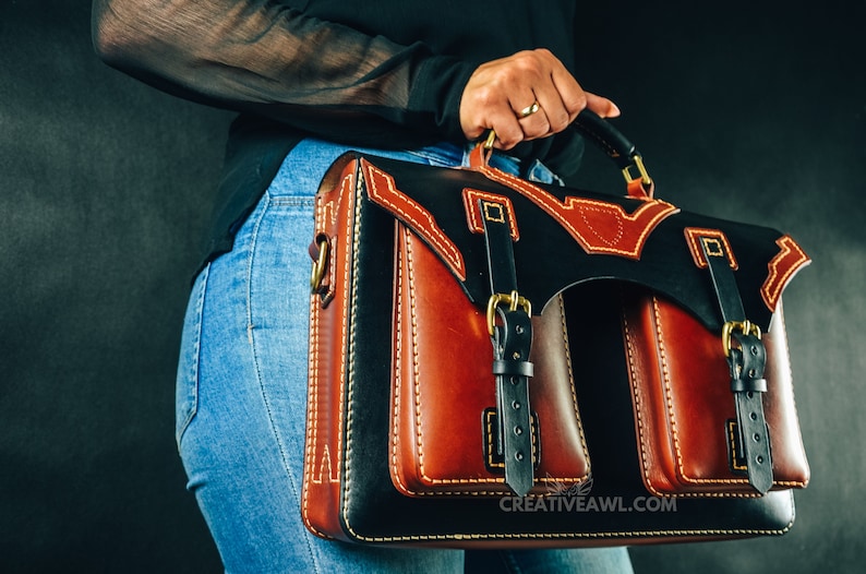 May include: A handcrafted leather briefcase with a striking design. The bag features a combination of black and brown leather, accented with cream stitching and gold-colored hardware. The bag has two front pockets and a top handle.