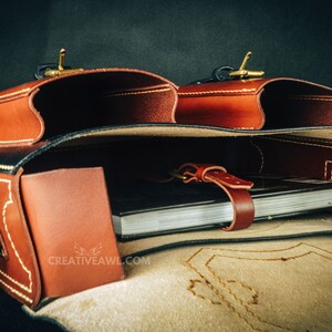 May include: A brown leather satchel with two exterior pockets and a flap closure. The bag features brass hardware and white stitching. Inside, a notebook is secured with a leather strap. The logo "CREATIVEAWL.COM" is visible.