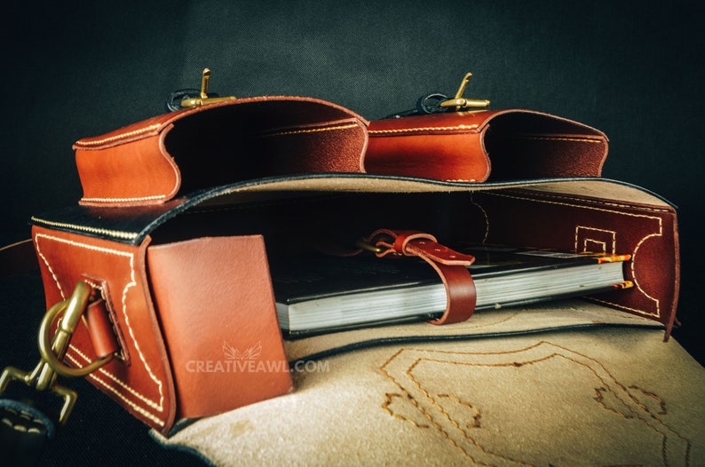 May include: A brown leather satchel with two exterior pockets and a flap closure. The bag features brass hardware and white stitching. Inside, a notebook is secured with a leather strap. The logo "CREATIVEAWL.COM" is visible.
