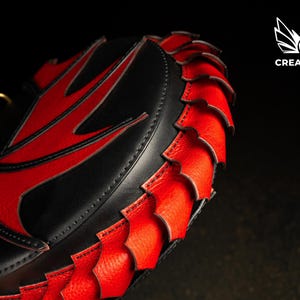 May include: A black and red leather bag with a distinctive design. The bag has a red accent on the top and a red, scale-like trim around the edge. A gold-coloured ring is attached to the top. The Creative Awl logo is in the upper right corner.