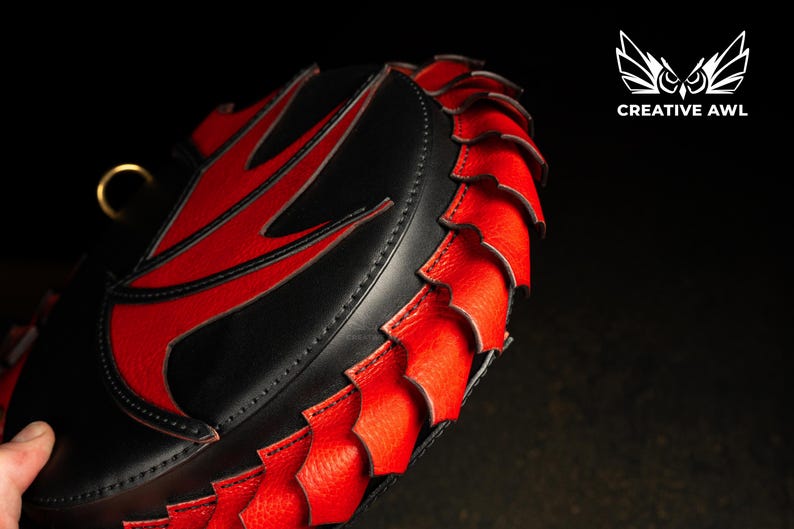 May include: A black and red leather bag with a distinctive design. The bag has a red accent on the top and a red, scale-like trim around the edge. A gold-coloured ring is attached to the top. The Creative Awl logo is in the upper right corner.