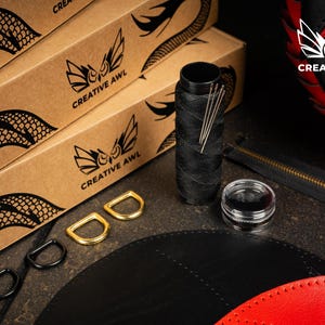 May include: Leathercraft supplies including black thread, needles, and D-rings. The image shows boxes with a dragon scale design and the "Creative Awl" logo, alongside a partially visible red and black leather item.