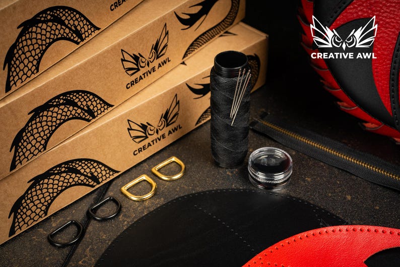May include: Leathercraft supplies including black thread, needles, and D-rings. The image shows boxes with a dragon scale design and the "Creative Awl" logo, alongside a partially visible red and black leather item.