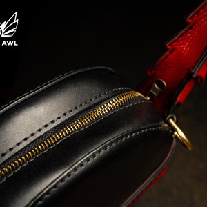 May include: Black leather bag with a gold zip and red strap. The bag has a rounded shape and is stitched with a contrasting thread. The strap is red with black accents. The bag has a gold-coloured zip.