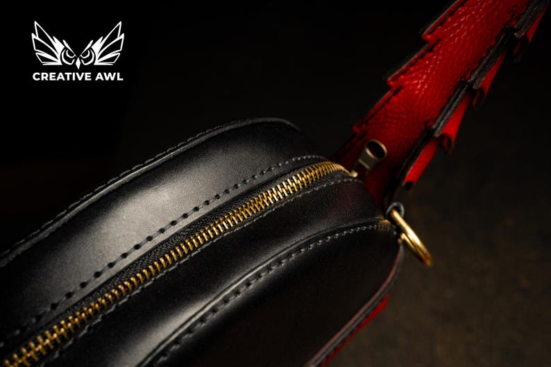 May include: Black leather bag with a gold zip and red strap. The bag has a rounded shape and is stitched with a contrasting thread. The strap is red with black accents. The bag has a gold-coloured zip.