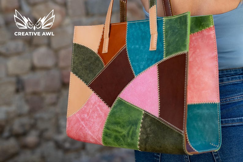 May include: A rectangular tote bag constructed from multi-coloured leather patches in shades of pink, green, blue, brown, and tan. The panels are joined with visible white cross-stitching. It features two light tan leather handles at the top.