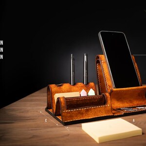May include: A brown leather desk organizer with a phone holder, pen holders, and a notepad section. The organizer is 8.9 inches wide, 5.3 inches high, and 5.3 inches deep. Two pens are in the pen holders.