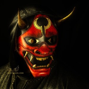 K&ouml;nnte beinhalten: Eine rote Leder-Oni-Maske mit goldenen Akzenten und schwarzem Hintergrund. Die Maske hat einen grimmigen Ausdruck mit scharfen Z&auml;hnen und H&ouml;rnern.