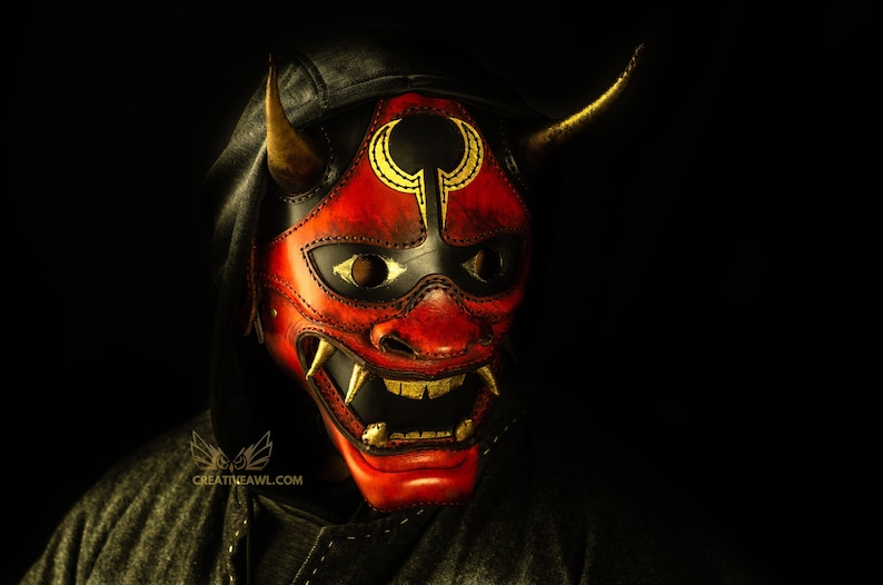 K&ouml;nnte beinhalten: Eine rote Leder-Oni-Maske mit goldenen Akzenten und schwarzem Hintergrund. Die Maske hat einen grimmigen Ausdruck mit scharfen Z&auml;hnen und H&ouml;rnern.