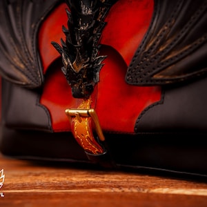 Op de afbeelding: Een zwarte leren tas met een drakenontwerp en een gouden gesp. De tas heeft een rood lederen paneel met een drakenontwerp.