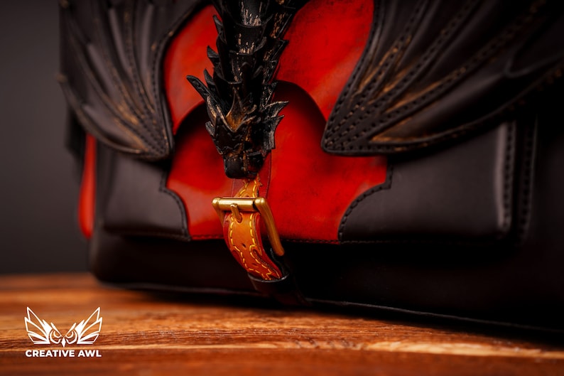 Op de afbeelding: Een zwarte leren tas met een drakenontwerp en een gouden gesp. De tas heeft een rood lederen paneel met een drakenontwerp.