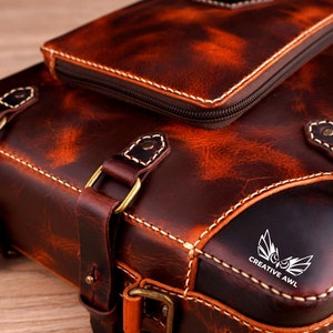 May include: A brown leather backpack with a front pocket and a logo that says "Creative Awl". The backpack has a distressed look and is made of high-quality leather.