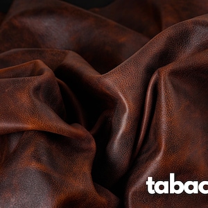 May include: Close-up of a brown leather fabric with a textured surface. The fabric is folded and draped, creating a soft, luxurious look. The word "tabaco" is printed in white on the fabric.