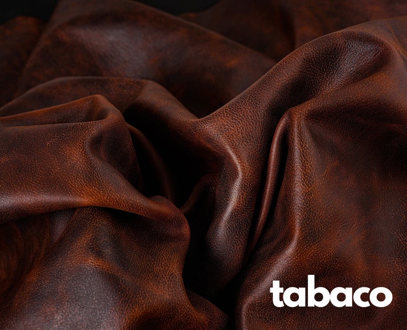 May include: Close-up of a brown leather fabric with a textured surface. The fabric is folded and draped, creating a soft, luxurious look. The word "tabaco" is printed in white on the fabric.