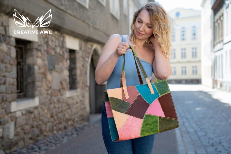May include: A rectangular tote bag constructed from multi-coloured leather patches in shades of teal, dark brown, olive green, light pink, and beige. The bag features visible stitching along the seams and two light tan leather shoulder straps.