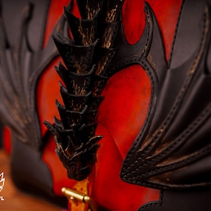 Op de afbeelding: Een rode leren tas met een zwart drakenontwerp. De vleugels van de draak zijn uitgespreid en zijn staart is opgerold. De tas heeft een gouden metalen sluiting.