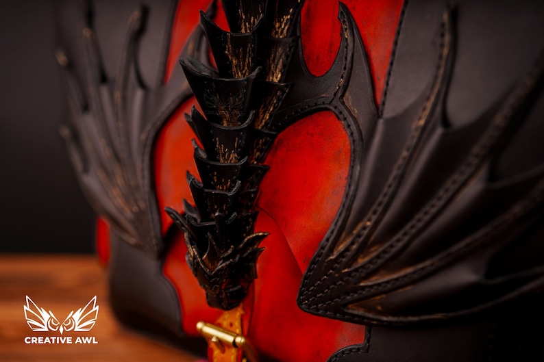 Op de afbeelding: Een rode leren tas met een zwart drakenontwerp. De vleugels van de draak zijn uitgespreid en zijn staart is opgerold. De tas heeft een gouden metalen sluiting.