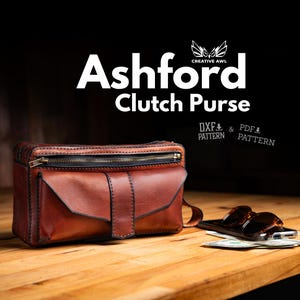 May include: A brown leather clutch purse with a zip and a flap pocket. The purse is on a wooden surface with sunglasses and money. The text "Ashford Clutch Purse" is displayed above the purse.