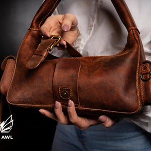 Puede incluir: Un bolso de mano de cuero marr&oacute;n con cierre de solapa y hebilla. El bolso tiene un bolsillo peque&ntilde;o en la parte delantera y un bolsillo m&aacute;s grande en la parte trasera. El bolso est&aacute; sostenido por la mano de una persona.